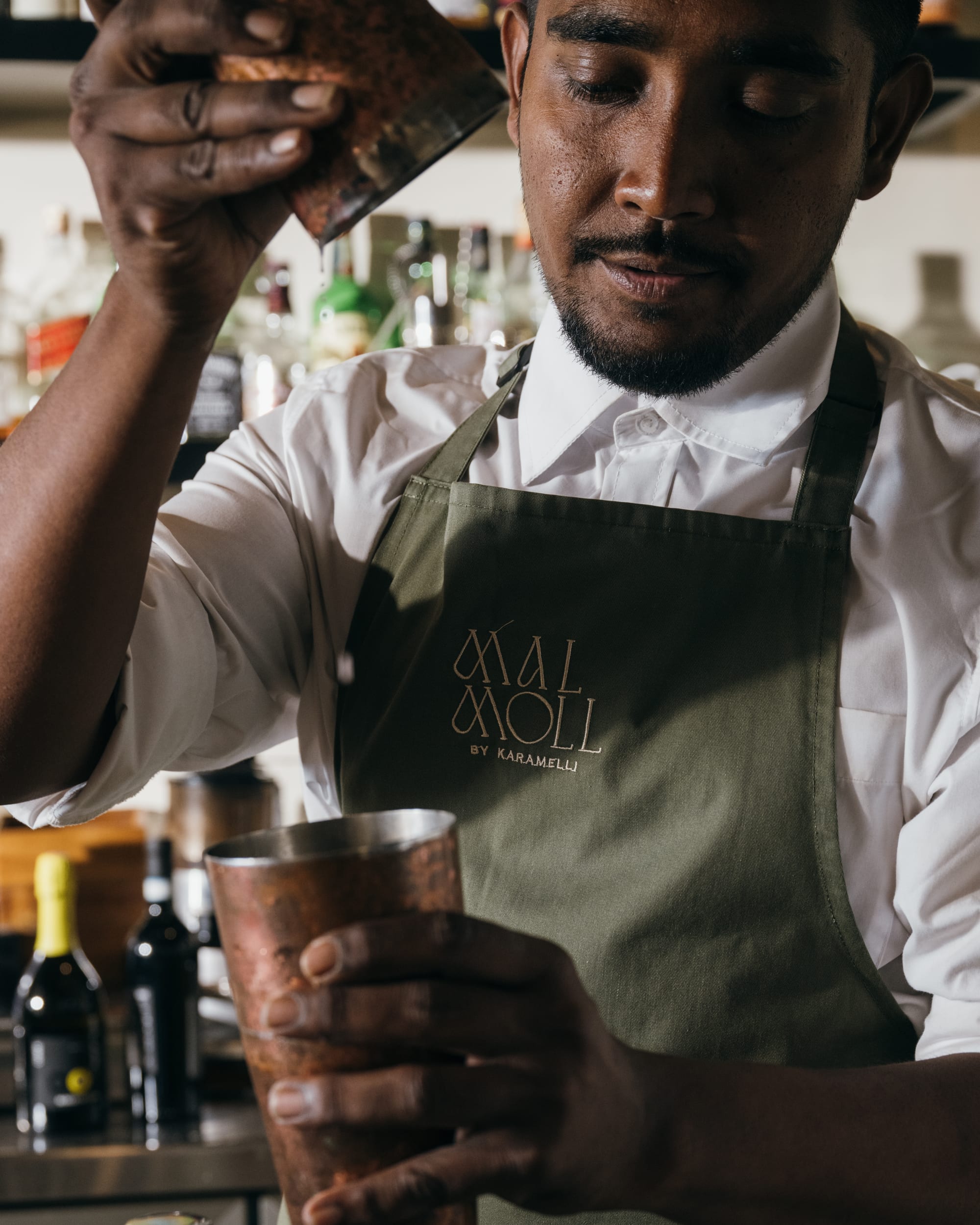 A bartender straining a cocktail, wearing the Mal Moll apron